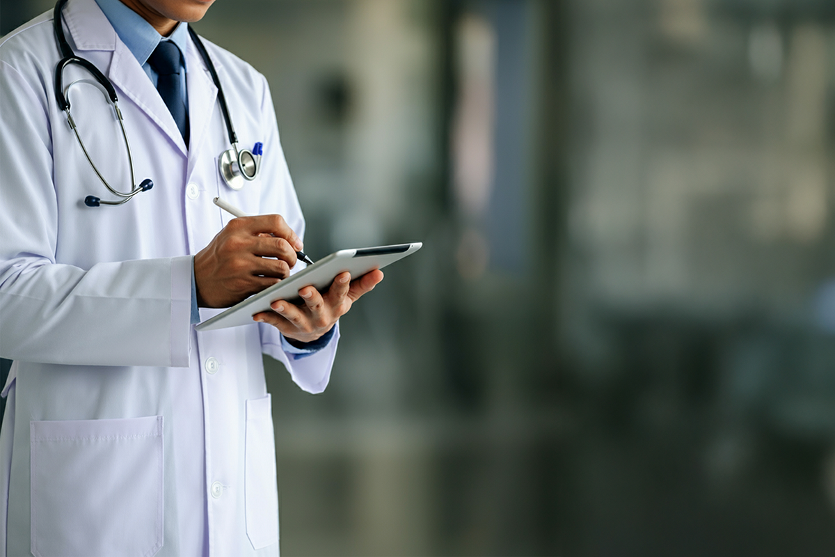 A medical doctor in a white coat with a stethoscope around his neck is writing something on the tablet in his hand